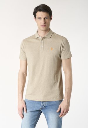 Giovane uomo che indossa una polo beige a maniche corte con piccolo logo arancione e jeans azzurri, in piedi davanti a uno sfondo uniforme.