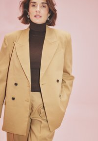 Woman with short brown hair wearing tan double-breasted suit and dark brown turtleneck against a pink background, one hand in pocket.