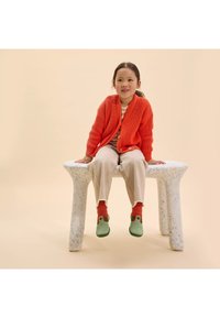 Dziecko siedzi na białej ławce w kropki z zaokrąglonymi nogami, ubranym w jasnopomarańczowy kardigan, beżowe spodnie i zielone buty z czerwonymi akcentami.