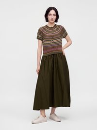 Femme debout avec une main sur la hanche, portant un pull à manches courtes à motifs, une longue jupe vert foncé et des baskets rose clair sur fond blanc.