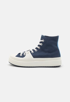 Marineblauwe hoge sneaker met witte rubberen zool en neuzenkap, witte veters en blauwe hielsluiting, getoond in zijaanzicht.