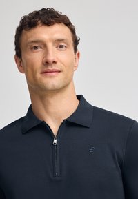 Marinblå zip-off pikétröja med krage, med en subtil logo på bröstet och en mjuk textur. Dragkedjan är silverfärgad med ett rundat drag.