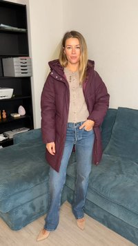 Tumši sarkans pūkains mētelis ar kapuci pār bēša krāsas pogājamu džemperi, kombinēts ar gaiši zilām izplatītām džinsu biksēm un bēša krāsas šauriem papēžiem.