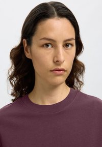 T-shirt cotone viola concollo a girocollo, con maniche corte e una superficie liscia, indossato da una persona con capelli ondulati di lunghezza media.