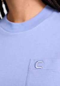 Maglietta di cotone azzurro chiaro con scollo tondo, maniche corte e una piccola lettera "C" ricamata sulla tasca frontale sinistra.