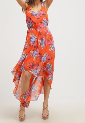 Frau mit einem orangefarbenen, ärmellosen Blumenkleid mit einem High-Low-Saum und beigen Plateausandalen, stehend mit erhobenen Armen.