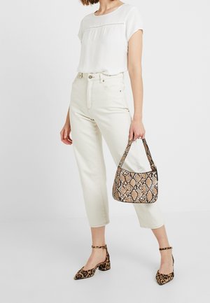 Eine Frau trägt ein weißes Oberteil und beige, gekürzte Jeans und hält eine Handtasche mit Schlangenledermuster. Sie kombiniert dies mit Leopardenmuster-Pumps.