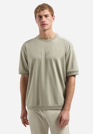 Man met licht beige korte mouw sweatshirt met de tekst "N.X.S COMFORT DELIVERY MOMENT", gecombineerd met bijpassende beige broek, staand frontaal.
