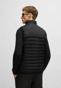 Schwarze gepolsterte Weste mit vertikalem Steppmuster, hohem Kragen und taillierter Passform. Getragen über einem schwarzen Langarmshirt mit minimalem Zubehör.