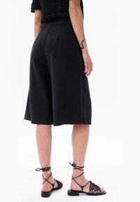 Zwarte culottes met een hoge taille, met een gladde textuur, losse pasvorm en knielange snit. Gecombineerd met zwarte vetersandalen.