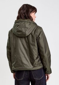 Veste matelassée vert olive avec capuche, présentant une texture lisse et des poignets élastiques. La veste a un design court, des coutures visibles et une capuche doublée de fourrure.