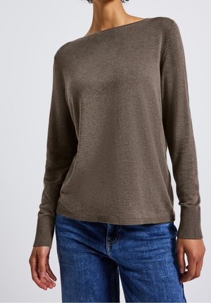 Femme portant un pull en maille marron à manches longues et un jean bleu en denim, debout devant un fond clair uni.