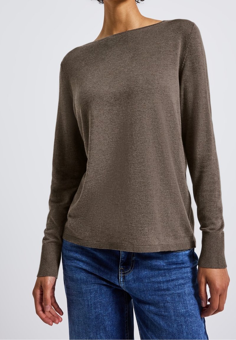 Femme portant un pull en maille marron à manches longues et un jean bleu en denim, debout devant un fond clair uni.