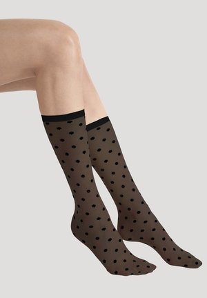 Beine, die durchsichtige schwarze Socken bis zur Mitte der Waden tragen, mit kleinen schwarzen Blumenmustern und einem einfarbigen schwarzen Bündchen vor neutralem Hintergrund.