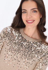 Beige pulóver bordás textúrával, ezüst és fekete flitterekkel díszítve, amelyek a felső részén színátmenetes mintában vannak elhelyezve.