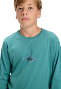 Türkisfarbene Langarm-T-Shirt aus Baumwolle. Mit einem diamantförmigen Logo in Grüntönen und Pink auf der Brust. Glatte Textur.