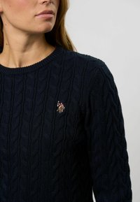 Námornícky pletený sveter s káblovým vzorom, okrúhlym výstrihom a vyšitým logom na hrudi. Mäkko textúrovaná tkanina a priliehavý strih.
