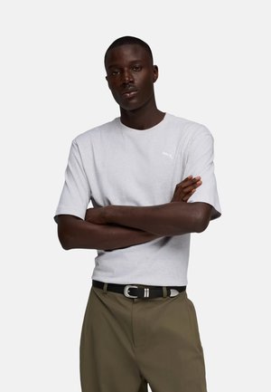 Uomo con le braccia incrociate che indossa una maglietta Puma grigio chiaro e pantaloni color oliva con una cintura nera su uno sfondo bianco semplice.