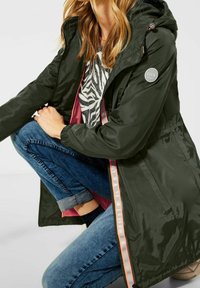 Olijfgroene waterdichte jas met capuchon, roze voering aan de binnenkant en contrasterende ritsdetails. Gecombineerd met een blauwe spijkerbroek en sokken.