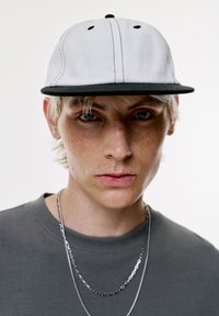 Fehér sapka fekete peremmel, fekete varrással és gomb részlettel, szürke póló fölött viselve, ezüstláncokkal rétegezve.