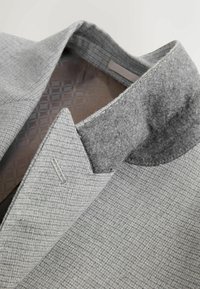 Grauer Blazer aus strukturiertem Stoff, mit einem gefalteten Kragen in dunklerem Grauton und dezentem Hahnentrittmuster.