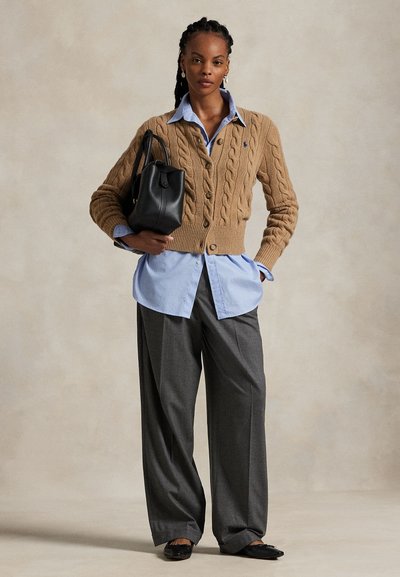 Femeie cu părul împletit poartă un cardigan bej tricotat cu model cu torsade peste o cămașă albastră, pantaloni largi gri, balerini negri și ține o geantă neagră din piele.