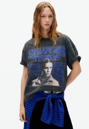Šedé nadrozmerné tričko s modrým grafickým motívom "Stranger Things", zavinuté rukávy a okrúhly výstrih. Textúrovaná látka s voľným strihom.