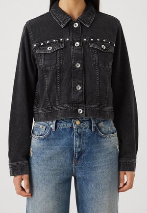 Giacca corta di jeans nera con bottoni e borchie argentate indossata con jeans blu sbiaditi a vita alta.