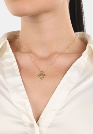 Collar con colgante de corazón dorado sobre una cadena delicada, sobre una blusa suave de color crema con un ligero brillo y diseño de escote en V.