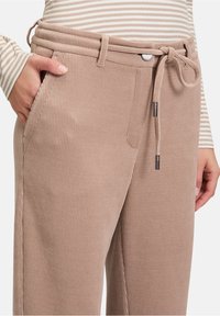 Beżowe spodnie z kordurowej tkaniny z pasem ze sznurkiem, kieszeniami po bokach i subtelną teksturą prążkowaną; ozdobione srebrnym guzikiem i detalami metalowymi.