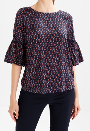 Marineblauwe blouse met rood bloemenpatroon en geplooide ellebooglange mouwen, gedragen met zwarte broek, getoond van nek tot heupen.