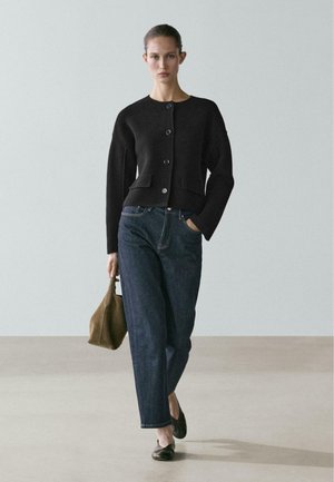 Donna che cammina in avanti indossando un cardigan nero con bottoni, jeans blu scuro, ballerine nere e tenendo una borsa marrone nella mano sinistra.