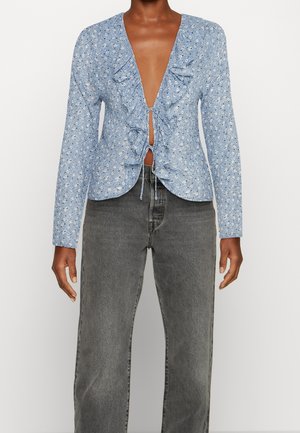 Blouse à manches longues à fleurs bleues avec bordure à volants et liens à l'avant, associée à un jean noir délavé taille haute sur une personne debout.