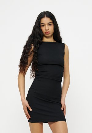 Vrouw met lang krullend haar, gekleed in een nauwsluitende, mouwloze zwarte jurk, staand tegen een effen lichte achtergrond.