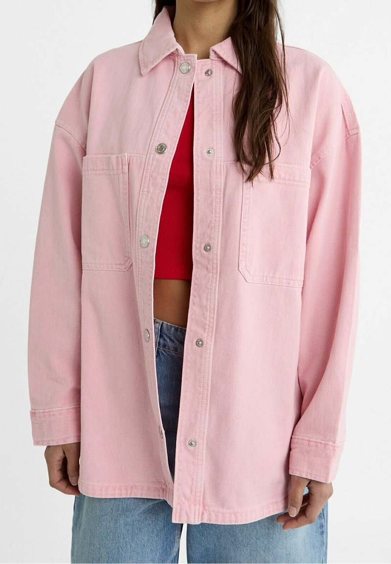 Chaqueta de mezclilla rosa con cierre de botones, cuello, bolsillo en el pecho y mangas largas. Llevada sobre una blusa roja con jeans azules.
