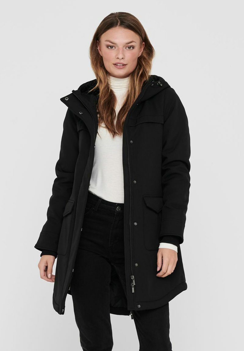 Parka noire, longue, avec une fermeture éclair et des boutons-pression, des poches avant, une capuche ajustable et une texture lisse. Portée par-dessus un pull léger.