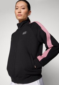 Zwarte ritsjack met roze zijstrepen, voorzien van een logo op de borst, twee zijzakken en een hoge kraag. Soepele stofstructuur.