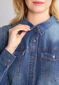 Denim-paita, jossa on napitettava kaulus, kahdella etupussilla ja hopeansävyisillä napeilla. Kankaan väri on haalistunut sininen.