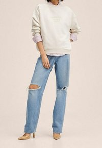 Hellgraues Sweatshirt mit dem Prägeaufschrift "MAJESTIC", geschichtet über einem pink gestreiften Hemd. Zerrissene blaue Jeans mit Rissen an den Knien, kombiniert mit beigen High Heels.