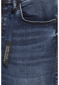 Blå denimjeans med en slät yta, med orange sömmar och en etikett som säger "KRÄV DIN DENIMRÄTTIGHET." Klassisk fem-fickorsdesign.