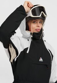 Chaqueta de esquí en blanco y negro con capucha, bolsillos con cremallera y puños ajustables. Presenta un parche con el logo en el frente y forro de malla.