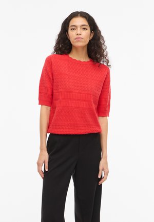 Femme aux longs cheveux bouclés portant un pull rouge texturé à manches courtes et un pantalon noir, debout devant un fond blanc.