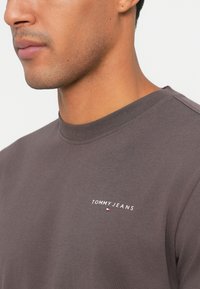 Zbliżenie na mężczyznę w brązowej koszulce z okrągłym dekoltem, z napisem "TOMMY JEANS" oraz małym logo flagi na piersi.