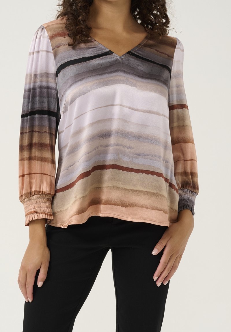 Femme portant un chemisier à manches longues avec des rayures horizontales aquarellées dans des tons bruns, gris et crème, associé à un pantalon noir.