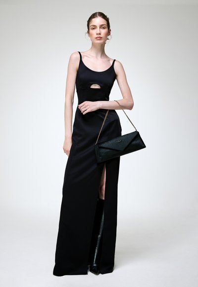 Femme portant une longue robe noire avec une fente à la cuisse et un décolleté découpé, tenant une pochette noire, debout devant un fond clair uni.
