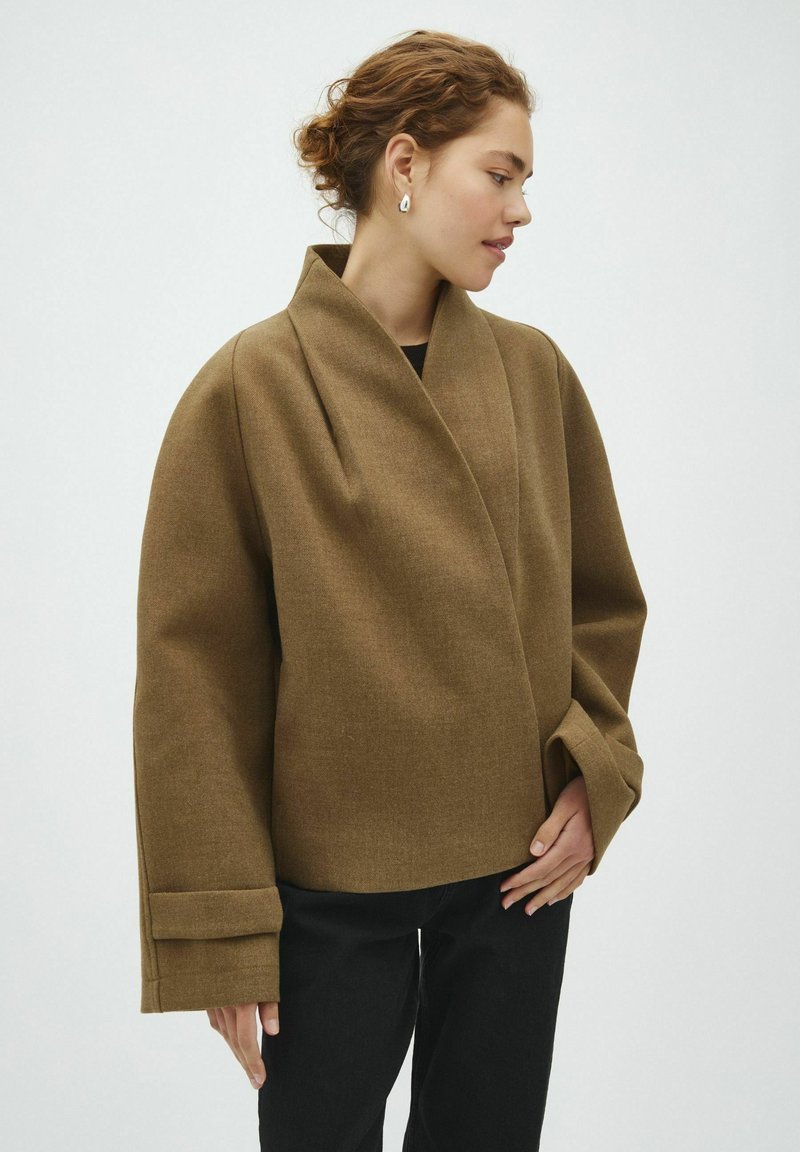 Femme aux cheveux bouclés porte un manteau marron oversized avec de larges revers et un pantalon noir, debout devant un fond clair uni.