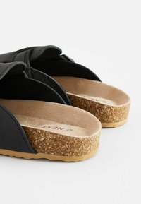 Svarta slip-in sandaler med korkfotbäddar och justerbara remmar på vit bakgrund.