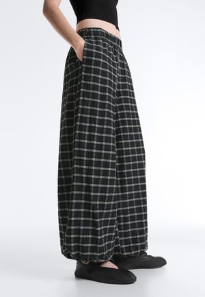 Personne portant un pantalon ample à carreaux noir et blanc avec une ceinture élastique et des chaussures noires slip-on, mains dans les poches, haut court noir.