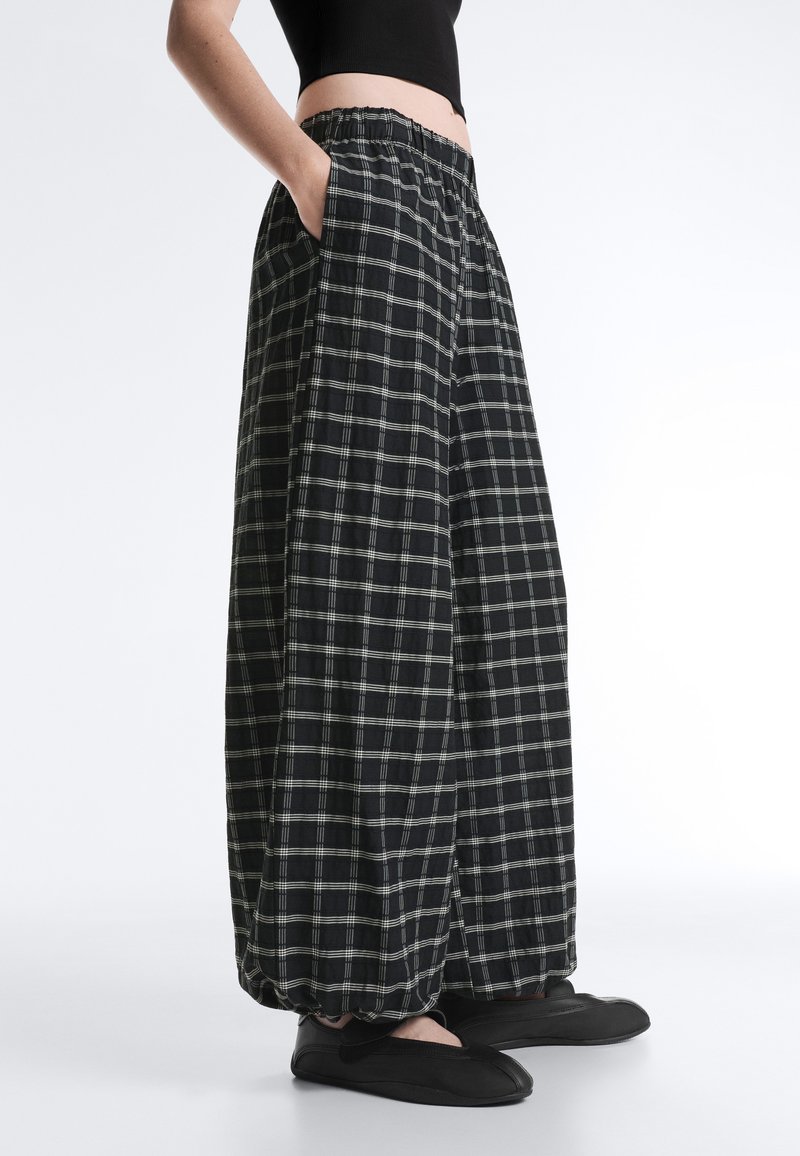 Personne portant un pantalon ample à carreaux noir et blanc avec une ceinture élastique et des chaussures noires slip-on, mains dans les poches, haut court noir.