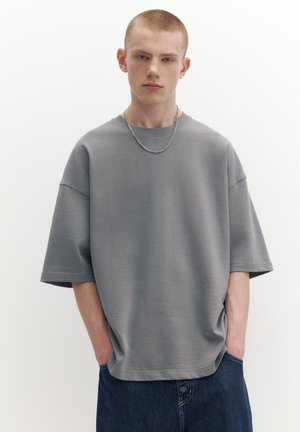 Mladý muž s krátkými vlasy, který nosí volné šedé tričko, stříbrný řetízek a tmavě modré džíny, stojí s rukama v kapsách.